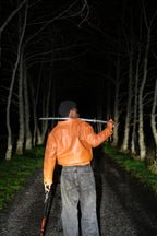 Person in a brown leather jacket holding a sword in a dark forest. Breastplate of Righteousness Leather Jacket in Brown. Back. Ephesians 6:14. LOVE GOD BIG Christian clothing brand streetwear faith-based apparel.