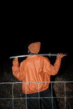 Person wearing a brown jacket holding a sword over their shoulder against a dark background. LOVE GOD BIG Christian clothing brand streetwear faith-based apparel.